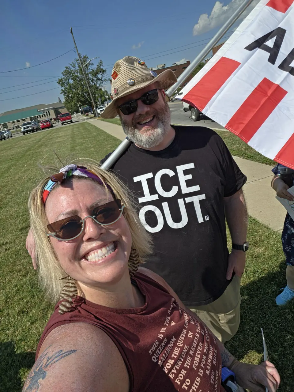 Two protesters smile at the camera on a grassy corner next to a road. One wears a black T-shirt that says ICE OUT and holds a pole with a red and white striped flag.